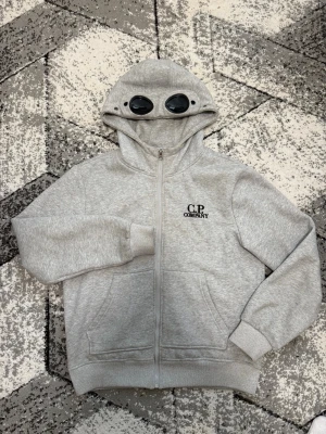 Grå zip hoodie med goggles C.P. Company - Cool grå hoodie från C.P. Company med zip och ikoniska goggles i huvan. Tröjan har broderad logga på bröstet, ribbade muddar och två stora fickor framtill. Perfekt för dig som gillar streetwear och unik design. Perfekt för sommarkväll !