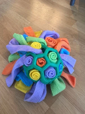 Färgglad aktiveringsboll för husdjur - En grön aktiveringsboll i gummi fylld med mjuka tygremsor i lila, orange, gul, blå och grön. Perfekt för att gömma godis och stimulera ditt husdjur mentalt. Bollen har ett nätmönster och tygremsorna är rullade och sticker ut ur öppningarna.