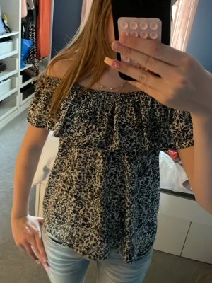 Svartvit blommig off shoulder topp - Snygg off shoulder topp med svartvitt blommigt mönster och volang upptill. Perfekt till jeans eller kjol för en trendig look. Lätt och luftig modell som sitter bekvämt över axlarna.