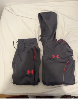 Under Armour mörkgrå träningsset - Mörkgrått träningsset från Under Armour med röda detaljer. Setet består av en hoodie med dragkedja och matchande byxor med resår i midjan och snörning. Båda plaggen har Under Armour-loggan framtill. Perfekt för träning eller chill.