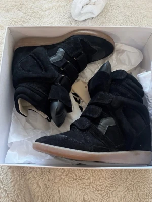 Svarta Isabel Marant wedge sneakers - Säljer ett par svarta wedge sneakers från Isabel Marant med högt skaft och kardborreband. Skorna har en blandning av mocka och läderdetaljer samt en beige sula. Perfekta för dig som vill ha en edgy och trendig look.