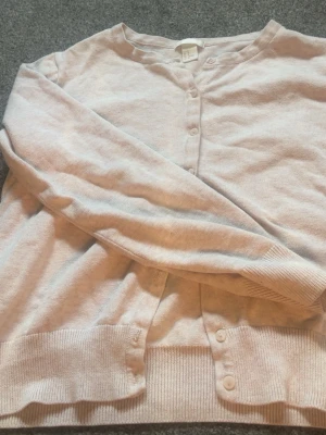Ljusbeige kofta från H&M - En stilren ljusbeige kofta från H&M med rund halsringning och knäppning framtill. Koftan har långa ärmar och ribbade muddar vid ärmslut och nederkant. Perfekt att slänga över en t-shirt eller linne för en chill look. Det står att den är i storlek M men passar mer som S