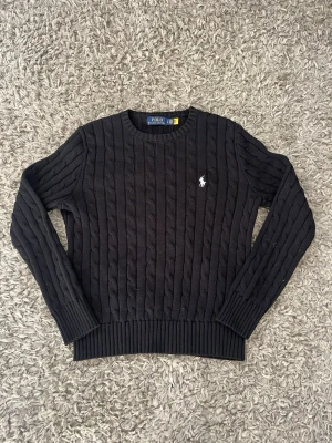 Svart kabelstickad tröja Polo Ralph Lauren - Kabelstickad svart tröja från Polo Ralph Lauren med rund halsringning och den klassiska vita logon broderad på bröstet. Tröjan har långa ärmar och ribbade muddar vid ärmslut och nederkant. Perfekt för dig som gillar stilrena och tidlösa plagg.