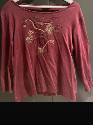 Vinröd tröja med broderade blommor - Säljer en vinröd tröja med rund halsringning och broderade blommor i beige och rosa på bröstet. Tröjan har långa ärmar och en avslappnad passform. Perfekt för dig som gillar detaljer och en romantisk stil. Den är i k men passar mer m eller s