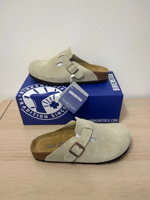  Birkenstock Boston Khaki  - Säljer ett par snygga beige Birkenstock Boston tofflor i Khaki med justerbart spänne i metall och klassisk korksula. Perfekta för dig som vill ha både stil och komfort. Ikonisk design och mjuk innersula för extra skön känsla.