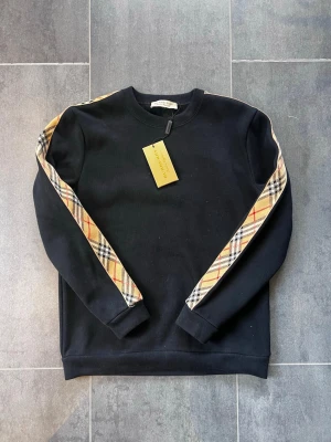 Svart sweatshirt från Burberry - Cool svart sweatshirt från Burberry med klassiskt rutmönster i beige, svart och rött längs ärmarna. Tröjan har rund halsringning och ribbade muddar. Perfekt för dig som gillar stilrena designer och ikoniska detaljer.