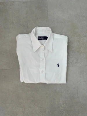 Vit skjorta från Polo Ralph Lauren - Klassisk vit skjorta från Polo Ralph Lauren med mörkblå broderad logga på bröstet. Skjortan har knappar framtill och en stilren krage. Tillverkad i bomull för en skön och luftig känsla.