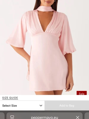 Ljusrosa kort klänning från PepperMayo - Söt ljusrosa kort klänning från PepperMayo med v-ringning, vida trekvartsärmar och matchande halsbandsscarf. Tillverkad i ett glansigt material som ger en elegant känsla. Perfekt för dig som vill sticka ut med en feminin och trendig look. Aldrig använd 