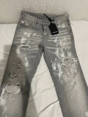 Grå slitna jeans från Dsquared2 - Säljer ett par grå jeans från Dsquared2 med coola slitningar och vita färgstänk. Jeansen har en rak passform, klassisk femficksdesign och är tillverkade i denim. Perfekta för dig som gillar en edgy och trendig stil.