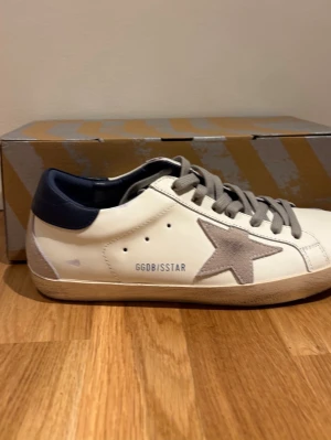 Golden Goose vita sneakers med stjärna - Säljer ett par  vita sneakers från Golden Goose med grå mockastjärna på sidan och grå skosnören. Skorna har en vintage-look med slitna detaljer och en brun sula i gummi. Perfekta för dig som gillar streetstyle och trendig design.
