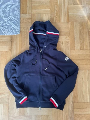 Mörkblå Moncler hoodie med dragkedja - Snygg mörkblå hoodie från Moncler med dragkedja och luva. Röd, vit och blå rand på huva och ärmslut. Klassisk Moncler-logga på ärmen och stora fickor framtill. Priset kan diskuteras. Skriv till mig för fler bilder 