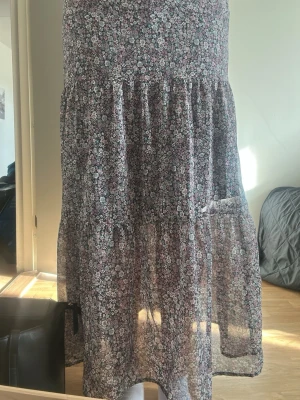 Blommig långkjol med volang - Säljer en luftig långkjol med småblommigt mönster i lila. Kjolen har volangdetaljer och är gjord i ett tunt, flowy material som passar perfekt till varmare dagar. Resår i midjan för skön passform.