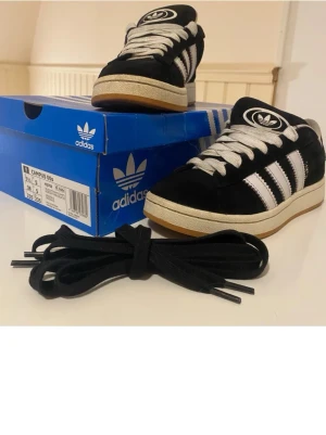 Adidas Campus 00s svart - Snygga Adidas Campus 00s sneakers i svart mocka med vita läderdetaljer och klassiska tre ränder på sidan. Skorna har vita tjocka snören, gummisula och Adidas logga på tungan. Perfekt för dig som gillar retrostil och streetwear.