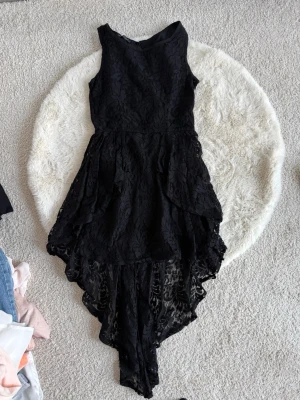 Svart asymmetrisk spetsklänning - Säljer en svart klänning med asymmetrisk kjol och vacker spetsdetalj. Klänningen är ärmlös och har en elegant, figurnära passform med unikt fall baktill som ger extra edge till looken.