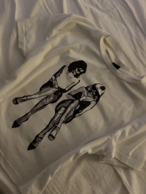 Vit t-shirt  - Vit t-shirt med ett stort svart grafiskt tryck av två sittande personer på framsidan. Klassisk rund hals och korta ärmar. Perfekt för dig som gillar unika motiv och streetstyle. Storlek XS/S