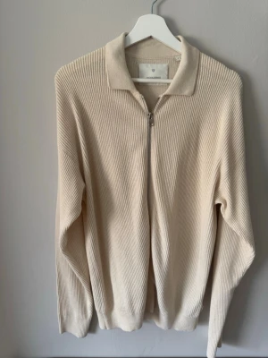 Beige stickad ziptröja från Jack & Jones  - Säljer en stilren beige stickad tröja från Jack & Jones med dragkedja framtill och klassisk krage. Tröjan har ribbad struktur, långa ärmar och är tillverkad i ett mjukt material som passar perfekt till lager på lager. Enkel och clean design. Storlek S men ganska stor i storlek.