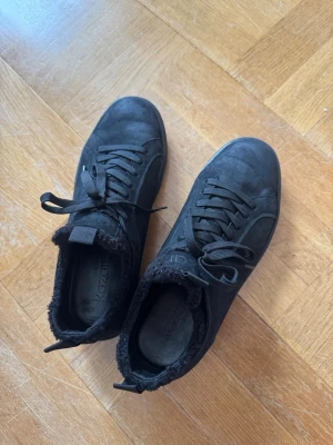 Svarta sneakers med fuskpälsfoder - Stilrena svarta sneakers från kazar med snörning och mjukt fuskpälsfoder runt kanten. Skorna har en klassisk låg siluett och ovandel i mockaliknande material. Perfekta för dig som vill ha både stil och komfort. Storlek 42 knappt använda.