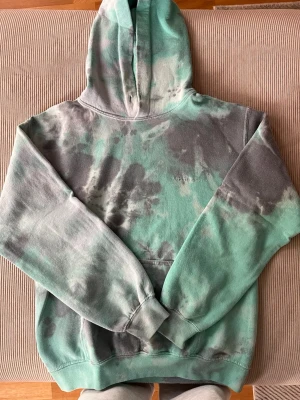 Grön och grå tie-dye hoodie - Cool hoodie med tie-dye mönster i grönt och grått från iets frans och urban outfitters. Den har en stor huva och broderad logga på bröstet. Perfekt för dig som gillar streetwear och vill sticka ut. Materialet är mjukt och bekvämt, med ribbade muddar och klassisk känguruficka.