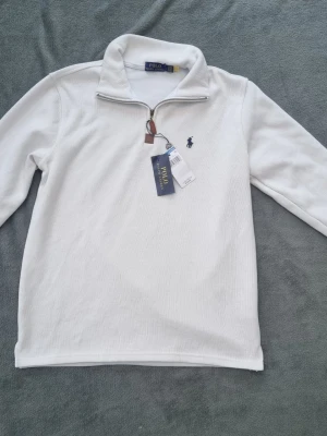 Beige quarter zip från Polo Ralph Lauren - Säljer en vit stickad tröja från Polo Ralph Lauren med halv dragkedja och klassisk broderad logga på bröstet. Tröjan har lång ärm och hög krage, perfekt för lager-på-lager. Materialet är mjukt och ribbstickat för extra komfort.