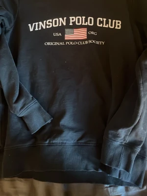 Svart sweatshirt Vinson Polo Club - Svart sweatshirt från Vinson Polo Club med tryckt logga och amerikansk flagga på bröstet. Tröjan har ribbade muddar vid ärmslut och nederkant samt rund halsringning. Perfekt för en sportig och avslappnad stil.