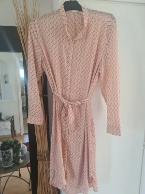 Ljusmönstrad omlottklänning med bälte - Säljer en ljus kortklänning med svart rutmönster och knytbälte i midjan. Klänningen har lång ärm, v-ringning och knappar framtill. Perfekt för dig som gillar en stilren och bekväm look.