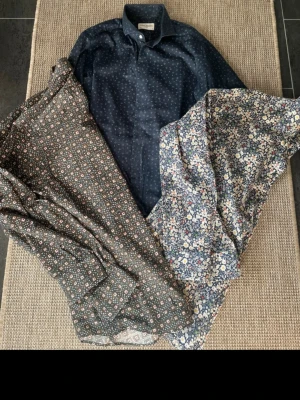 Mönstrade skjortor från John Henric - Tre skjortor i olika mönster från John Henric. En mörkblå skjorta med små vita prickar, en brun skjorta med blommigt mönster och en blå skjorta med blommor i vitt och rött. Alla har klassisk krage och knappar framtill. Perfekta för dig som gillar stilrena detaljer. En skjorta är i storlek 42