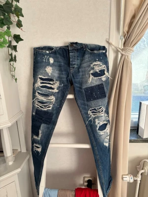Blå slitna jeans från Cipo & Baxx - Säljer ett par blå jeans från Cipo & Baxx med regular fit. Jeansen har stora slitningar, patchade detaljer och färgstänk framtill för en riktigt edgy look. Perfekta för dig som gillar streetstyle och vill sticka ut i mängden.