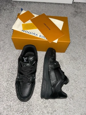 Louis Vuitton svarta sneakers - Snygga svarta sneakers från Louis Vuitton. Skorna har en låg siluett, är tillverkade i läder och har LV-logga på plösen. Perfekta för dig som vill ha något exklusivt och stilrent i garderoben.