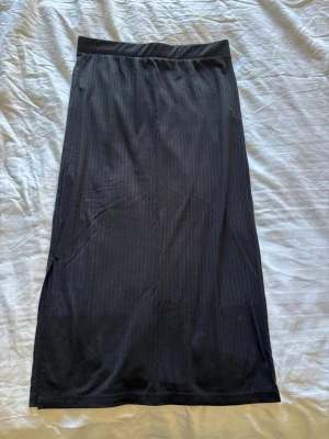 Svart ribbad långkjol med slits - Säljer en svart långkjol med diskret ribbat mönster och slits på sidan. Kjolen har en enkel och clean look som passar till det mesta. Materialet känns mjukt och stretchigt, perfekt för dig som gillar bekväma plagg.