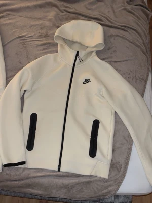 Nike tech fleece tracksuit i färgen beige - Säljer ett fräscht vitt set från Nike med matchande hoodie och byxor. Hoodien har dragkedja, huva och svarta detaljer vid fickorna samt Nike-logga på bröstet. Byxorna har dragsko i midjan, svarta fickor och Nike-logga. Perfekt för en sportig och avslappnad stil.