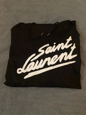 Saint laurent hoodie  - Svart hoodie från Saint Laurent med stor vit logga i graffiti-stil på bröstet. Klassisk huva med dragsko och magficka framtill. Tillverkad i mjukt material som känns skönt mot huden. Perfekt för dig som vill ha en streetwear vibe och exklusiv känsla. PRISET GÅR ATT  DISKUTERA!
