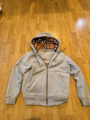 Burberry hoodie  - Snygg grå zip-hoodie från Burberry med klassiskt rutigt foder i luvan. Hoodien har dragkedja framtill, två fickor och ribbade muddar. Perfekt för dig som gillar stilrena plagg med en twist. Materialet är mjukt och bekvämt, troligen bomullsblandning.