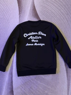 Svart sweatshirt  - Svart sweatshirt med vit broderad text på bröstet och ryggen. Tröjan har rund halsringning, långa ärmar och ribbade muddar vid ärmslut och nederkant. Perfekt för en stilren och lyxig look. Står storlek M men passar mer som S