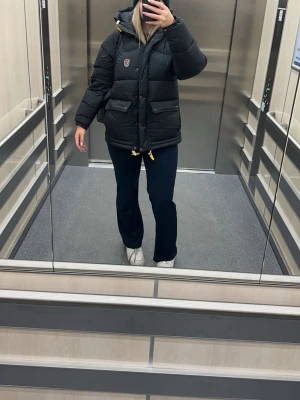 Svart dunjacka från Fjällräven - Säljer en svart, rejält vadderad dunjacka från Fjällräven med huva och gula detaljer. Jackan har två stora fickor framtill, dragsko i nederkant och klassisk Fjällräven-logga på bröstet. Perfekt för kalla vinterdagar.