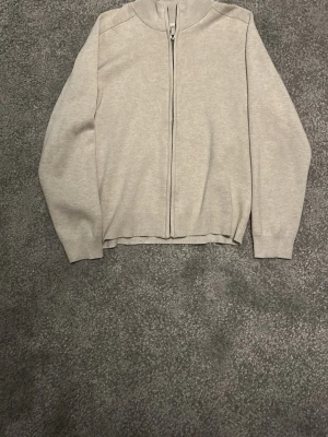 Beige ziptröja med hög krage - En stilren beige ziptröja med hög krage och hel dragkedja framtill. Tröjan har långa ärmar och ribbade muddar vid ärmslut och nederkant. Perfekt för lager-på-lager och enkel att matcha med olika outfits.