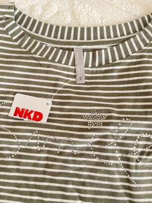 Randig t-shirt med pärlor från NKD - Snygg randig t-shirt från NKD i grönt och vitt med pärldetaljer på bröstet. T-shirten har korta ärmar och är gjord i mjuk bomull. Perfekt för dig som gillar en chill och trendig look med lite extra bling.