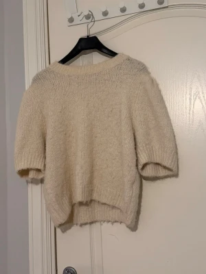 Krämvit stickad tröja med puffärm - Mysig krämvit stickad tröja med korta puffärmar och rund halsringning. Tröjan är stickad i ett mjukt, luddigt material som ger en trendig look. Perfekt att styla med jeans eller kjol för en chill vibe.