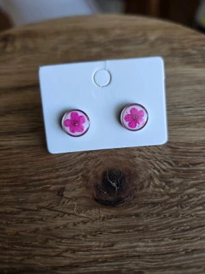 Runda örhängen med rosa blomma - Säljer ett par runda örhängen med motiv rosa blommor inuti på vit bakgrund. Örhängena är små och diskreta, perfekta för att ge en söt touch till din stil. Materialet är rostfritt stål.