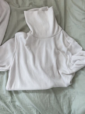 Vit ribbad polotröja - Säljer en vit ribbad polotröja med hög krage och långa ärmar. Tröjan är gjord i ett mjukt och stretchigt material som känns skönt mot huden. Perfekt för lager-på-lager och snygg att styla med jeans eller kjol.