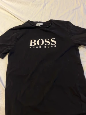 Svart Hugo Boss t-shirt - Snygg svart t-shirt från Hugo Boss med stor vit logga på bröstet. Klassisk rund halsringning och korta ärmar. Tillverkad i mjuk bomull som känns skön mot huden. Perfekt för dig som gillar stilrena och trendiga plagg. Pris kan diskuteras 