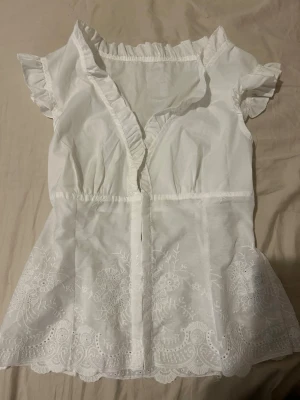 Vit blus med volang och brodyr - Söt vit blus med korta volangärmar och v-ringning med volangkant. Blusen har broderade detaljer nertill och lätt rynk vid midjan för en feminin siluett. Tillverkad i ett tunt, luftigt tyg som känns fräscht och somrigt.