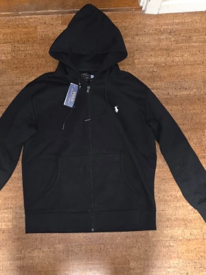 Svart hoodie från Polo Ralph Lauren - Svart hoodie med dragkedja från Polo Ralph Lauren. Klassisk design med vit broderad logga på bröstet, två fickor framtill och justerbar huva. Tillverkad i mjukt bomullsmaterial, perfekt för en avslappnad och stilren look.