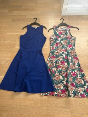 Blommig och blå 2 st kortklänning - Säljer 2 färgglad kortklänning med tropiskt blommönster i rosa, beige och grönt. Klänningen är ärmlös, har rund halsringning och en figurnära passform upptill med utställd kjol. Perfekt för sommarens alla tillfällen!