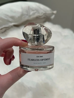 Fearless Optimist Eau de Toilette - En elegant parfymflaska i genomskinligt glas med en unik, vriden kork. Innehållet är ljusrosa och etiketten visar namnet Fearless Optimist. Parfymen är vegansk och Eau de Toilette, perfekt för dig som gillar fräscha och moderna dofter. Använd några gånger men fortfarande väldigt mycket kvar!