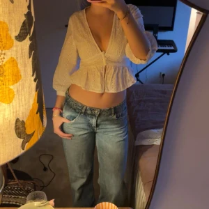 Vit broderad blus med v-ringning - Söt vit blus med broderade blommor, djup v-ringning och knappar framtill. Modellen har trekvartsärm och volang nedtill för en feminin look. Perfekt till jeans eller kjol för en trendig outfit.