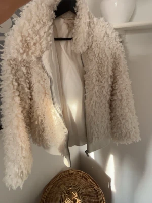 Krämvit teddyjacka med dragkedja - Supermjuk teddyjacka i krämvit färg från H&M. Jackan har en fluffig, lockig struktur och stängs med en silvrig dragkedja framtill. Kortare modell med hög krage som ger en trendig look. Perfekt för dig som vill ha en cozy vibe i höst och vinter.