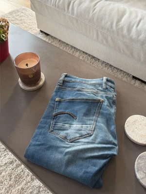 Blå slitna jeans från G-Star - Säljer ett par blå jeans från G-Star med slitningar och tvättade detaljer för en avslappnad look. Klassisk femficksmodell med raka ben och gylf med knapp. Perfekta för dig som gillar en casual och trendig stil. Använda men i väldigt fint skick! 