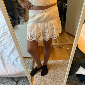 Vit kortkjol med broderade detaljer - Supersöt vit kortkjol från H&M med broderade blommönster och volangkant. Kjolen har resår i midjan och är gjord i lätt bomullstyg, perfekt för varma dagar. Kjolen har även inbyggda shorts under för extra komfort.