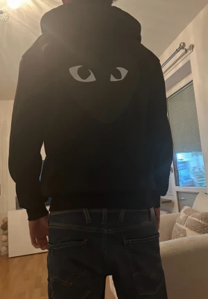 Svart hoodie med ögontryck - Svart hoodie från Comme des Garçons Play med ikoniskt hjärta med ögon-tryck på bröstet. Tröjan har huva med snörning, dragkedja framtill och två fickor. Materialet är mjuk bomull och passformen är normal. Perfekt för dig som gillar streetwear och stilrena detaljer.