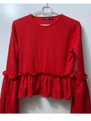 Röd volangtop från Boohoo - Trendig röd topp från Boohoo med volangdetaljer längs midjan och ärmarna. Modellen har rund halsringning och långa ärmar. Materialet är mjukt och stretchigt, perfekt för dig som vill sticka ut med en färgstark look.